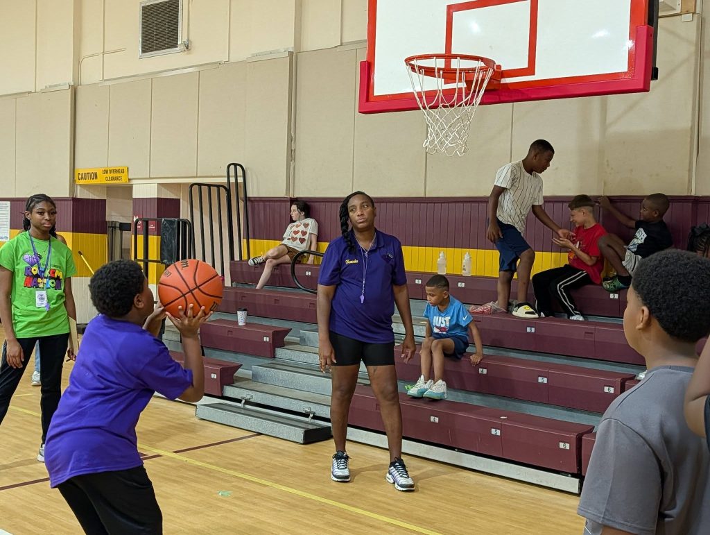 Children play basketball in a gymnasium as others sit on bleachers; two adults supervise. One boy prepares to shoot the ball near the hoop.