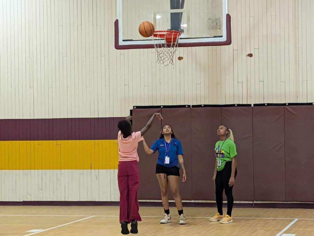 Three people in a gym watch as a basketball approaches the hoop; one person is actively shooting the ball.
