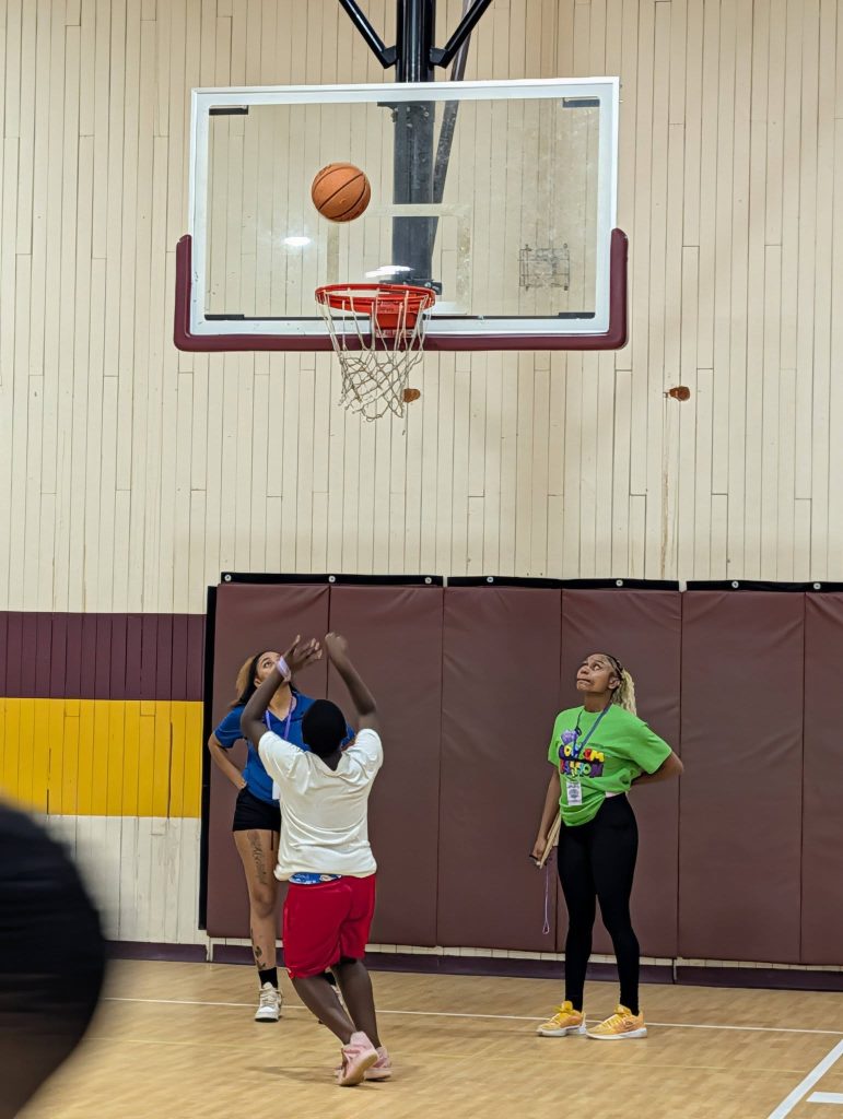 A child shoots a basketball towards the hoop in a gymnasium while two adults watch nearby.