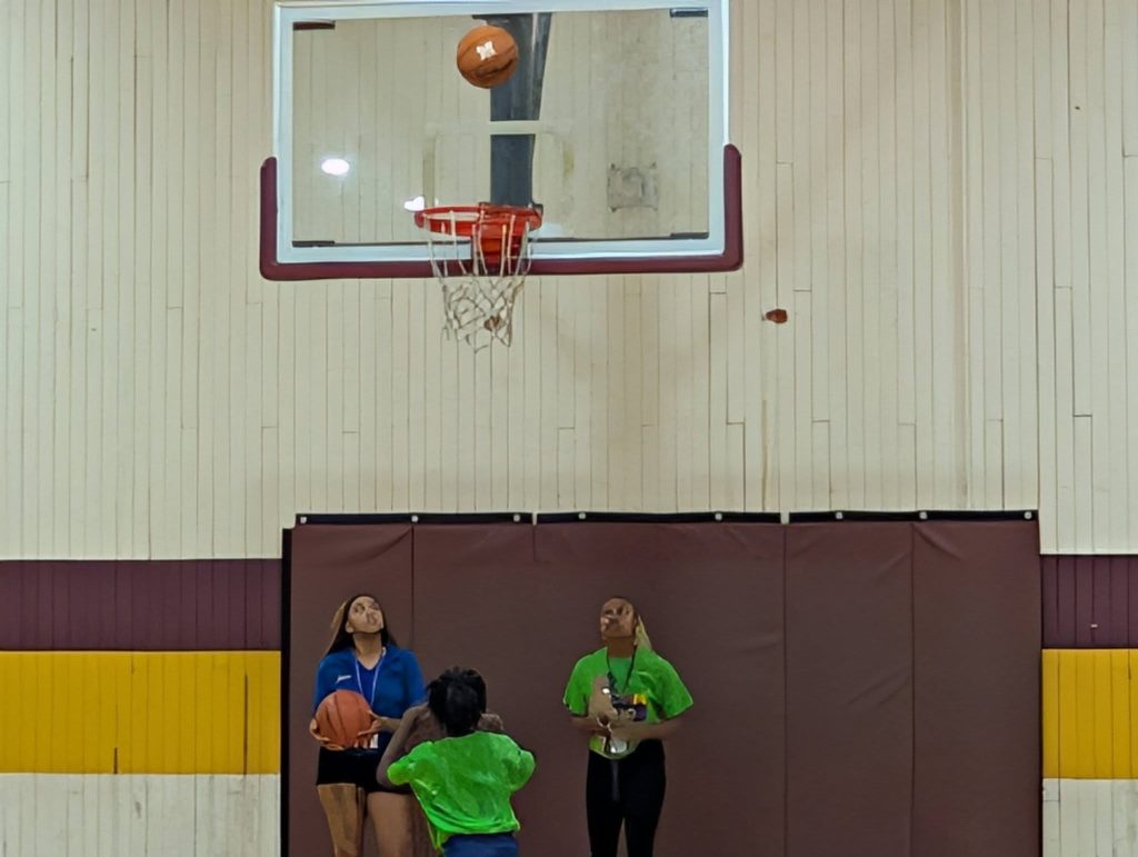 A child shoots a basketball towards the hoop in a gym while two adults watch, one holding another basketball.