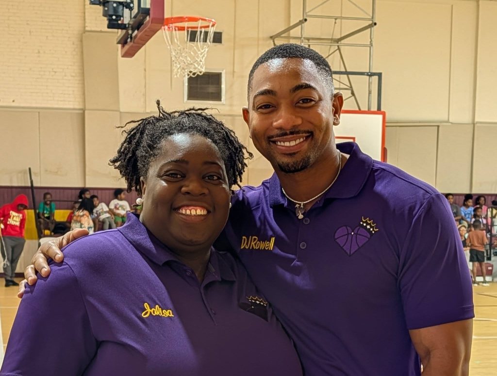 Two people smiling and posing together in a gym, both wearing purple shirts with embroidered names and a heart-basketball logo. Basketball hoop and people in the background.