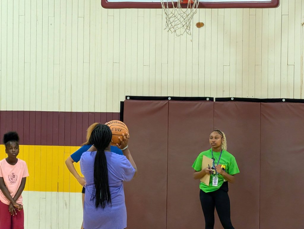 Three girls and a coach stand in a gym. One girl prepares to shoot a basketball while others observe.