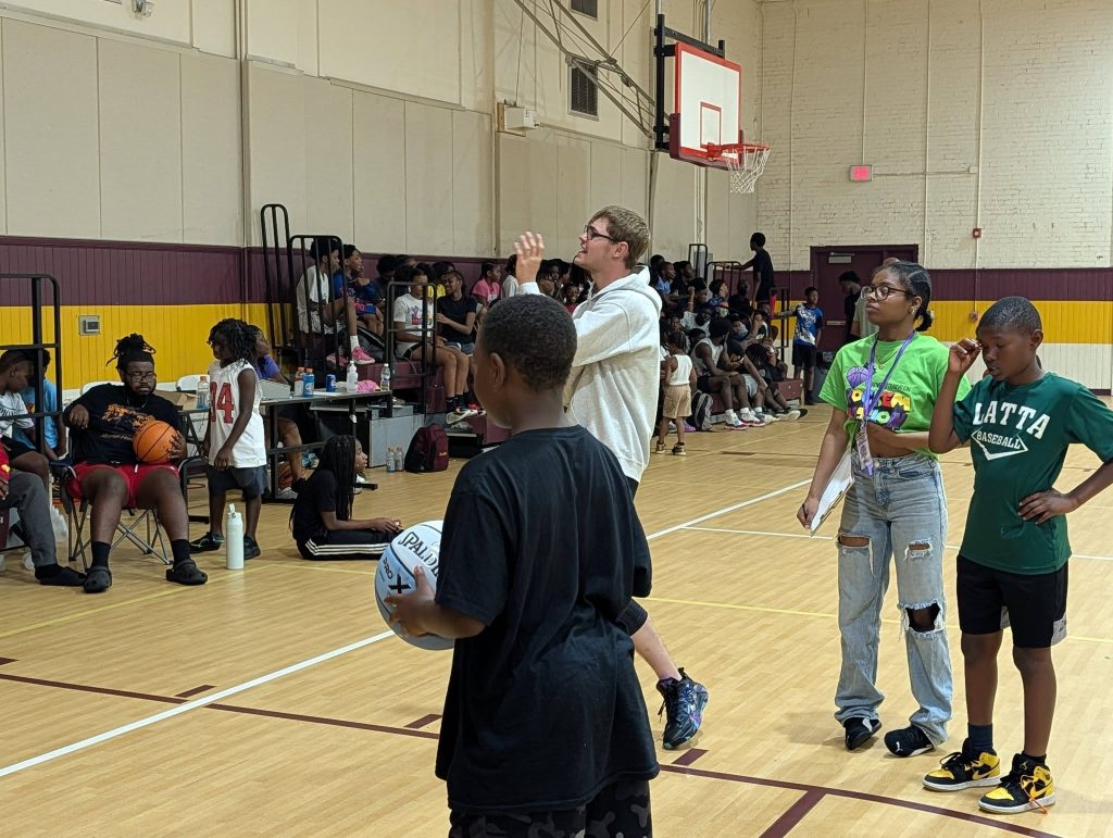A group of children and adults gather in a gymnasium, some holding basketballs, with spectators seated in the bleachers in the background.