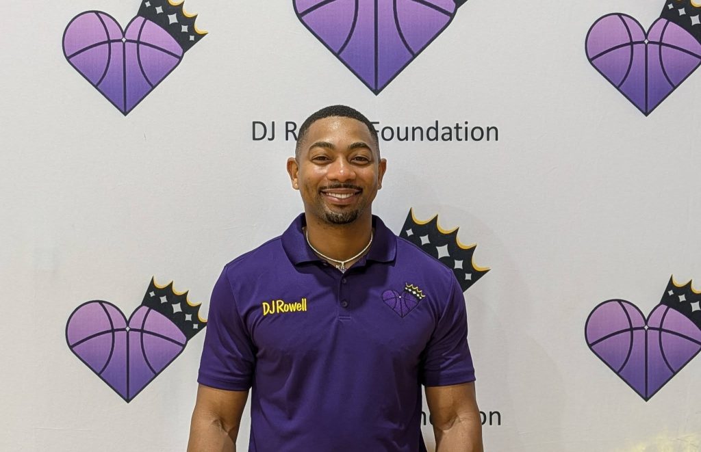 Smiling man in a purple DJ Rowell Foundation polo stands in front of a backdrop with purple heart basketball logos and crowns.