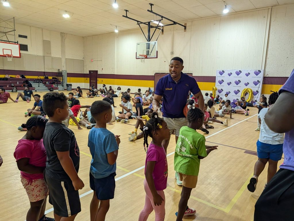 A group of children and an adult interact in a gymnasium with basketball hoops, surrounded by other seated kids and colorful decorations.