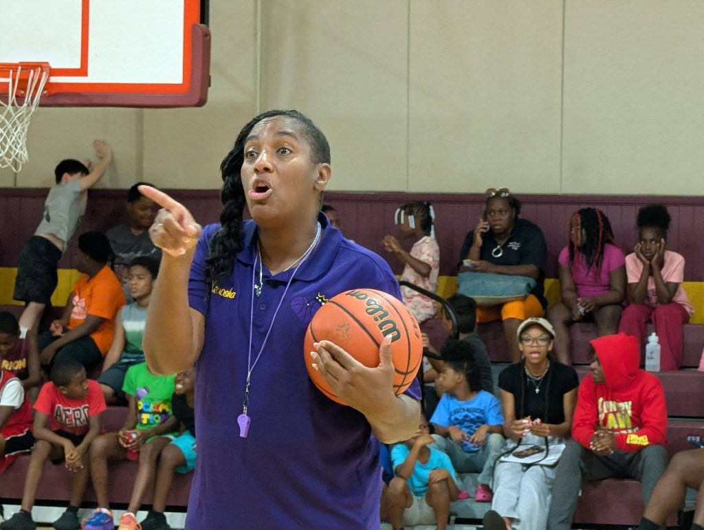 Female basketball coach holding a ball and instructing players in a gym, with children and adults seated in the bleachers behind her.