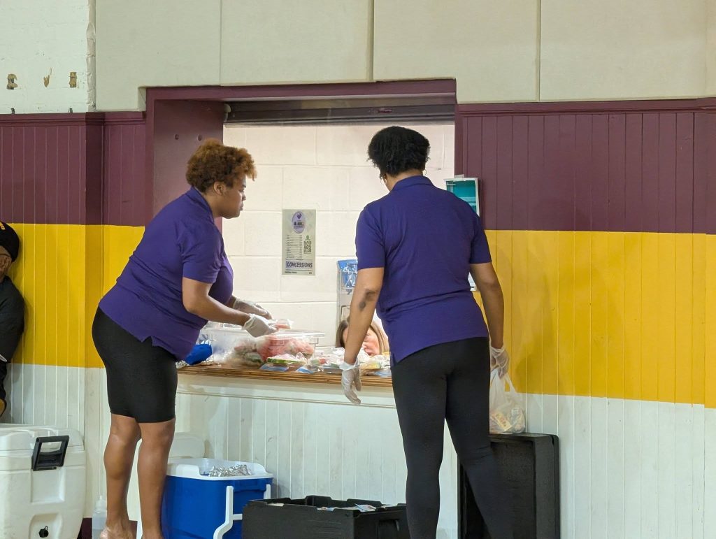 Two women in purple shirts serve food at a concession stand, wearing gloves, inside a building with maroon and yellow walls.