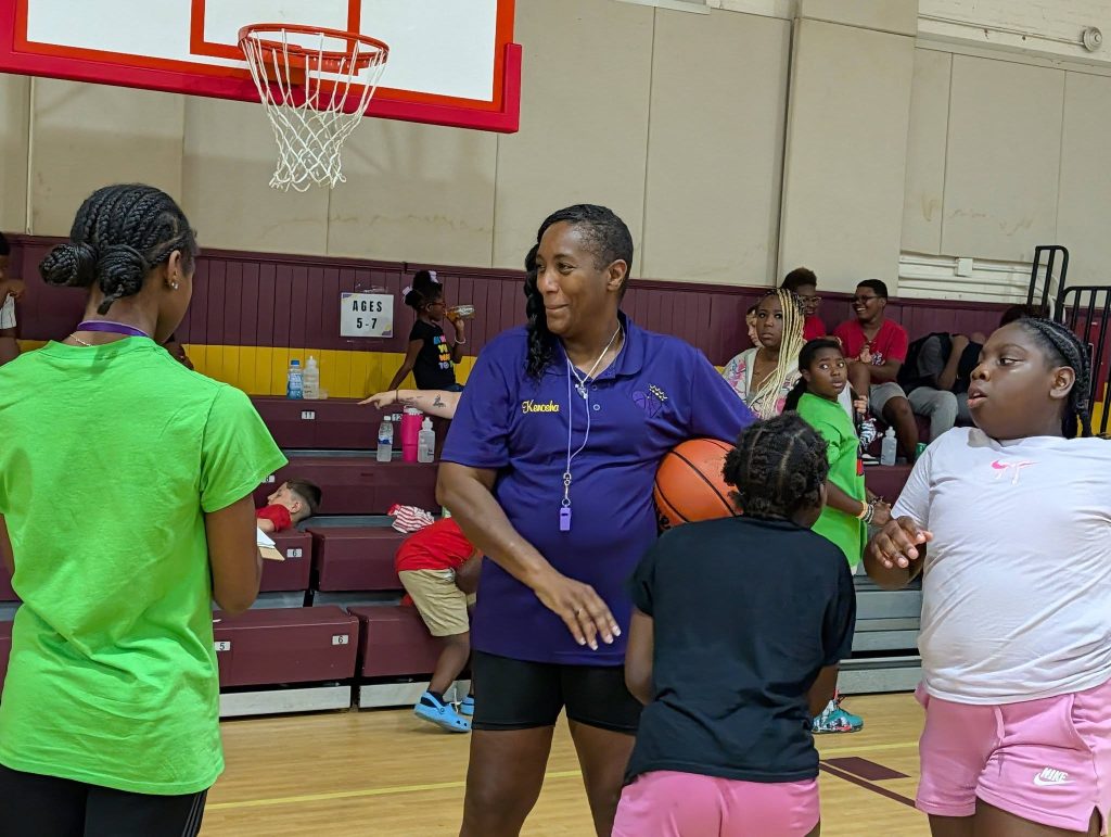 A basketball coach holding a ball talks to three young girls in a gym, with people sitting on bleachers in the background.