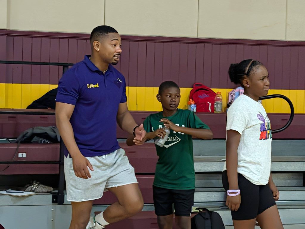 A man in a blue shirt talks to two kids in athletic clothes inside a gymnasium with benches and backpacks in the background.