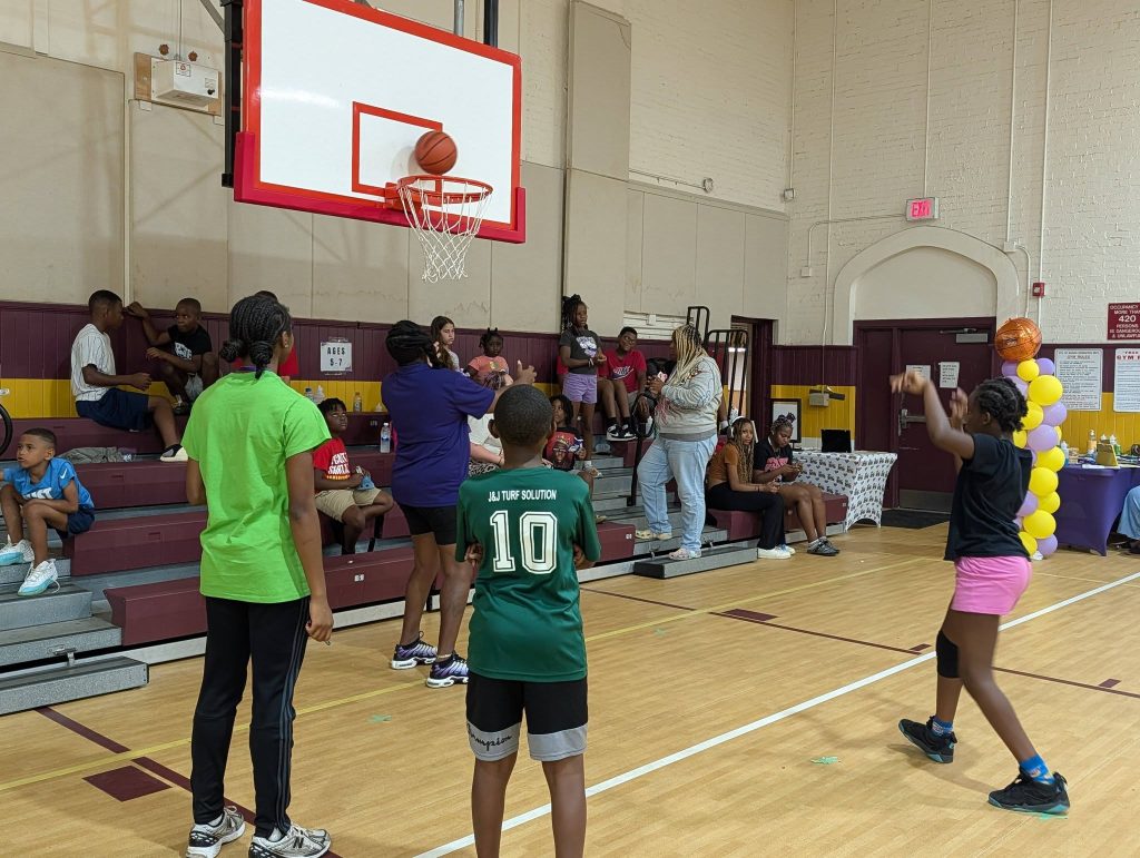 Children play basketball in a gym while others watch from the bleachers; a girl shoots the ball as it nears the hoop.