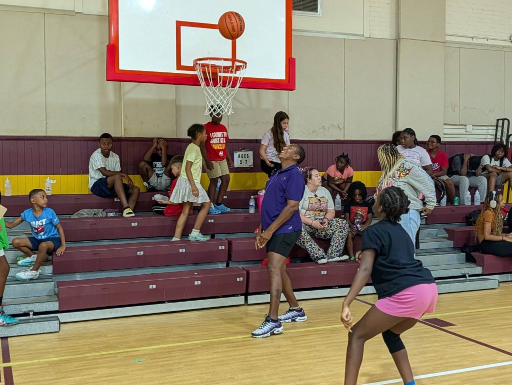 People play basketball in a gymnasium while others sit and watch on bleachers in the background.
