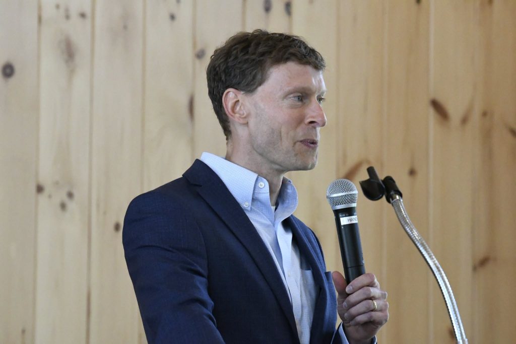 A man in a blue suit speaks into a microphone while standing indoors against a wooden wall background.