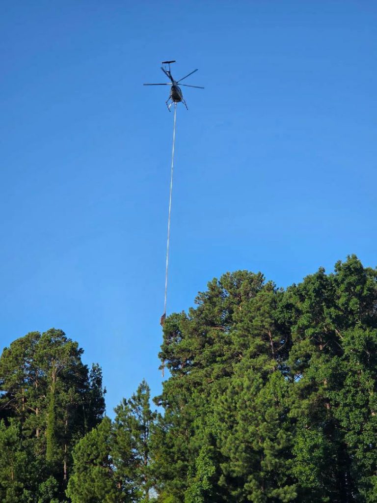 A helicopter hovers above a forest, carrying a load suspended by a long cable, with green trees below and clear blue sky above.