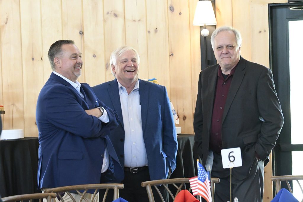 Three men in business attire stand and converse at an indoor event, with wooden walls and a table displaying small American flags and a table number six sign.