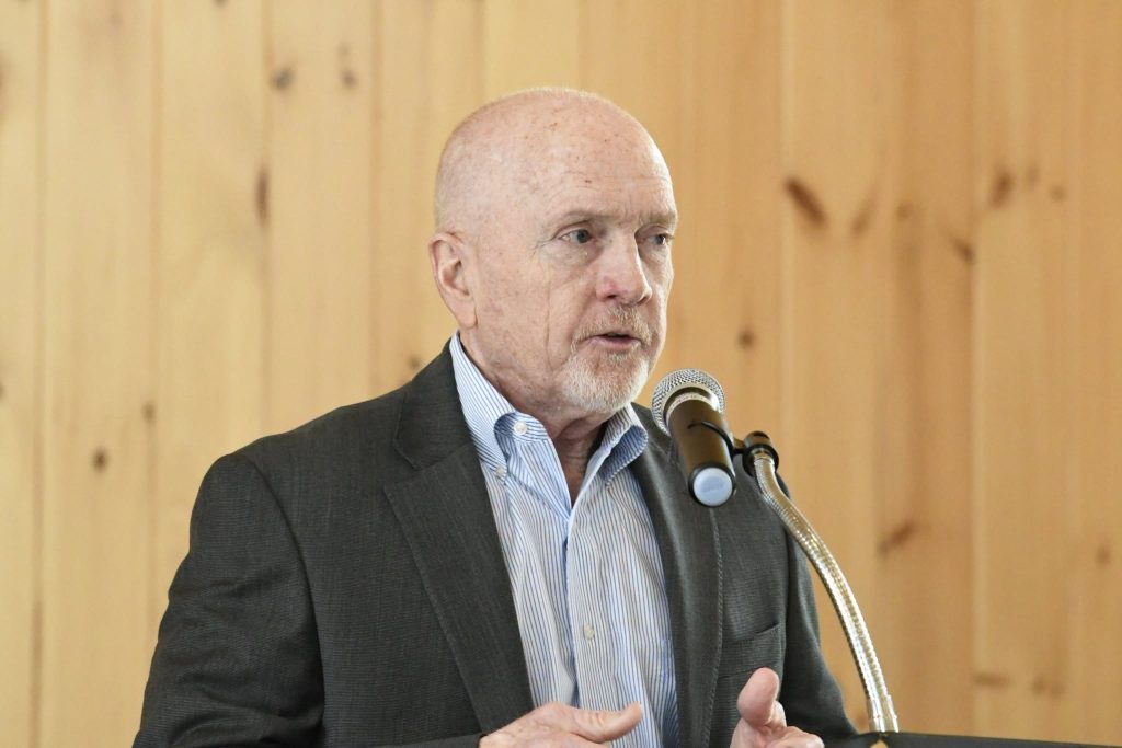 older man in a suit speaks into a microphone in front of a wooden wall, appearing to give a formal speech or presentation.