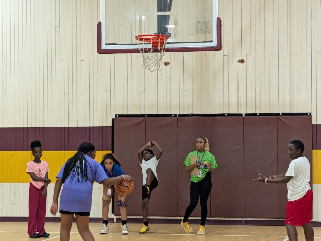 Six kids stand in a gymnasium under a basketball hoop, with one holding a basketball, ready to play or practice.