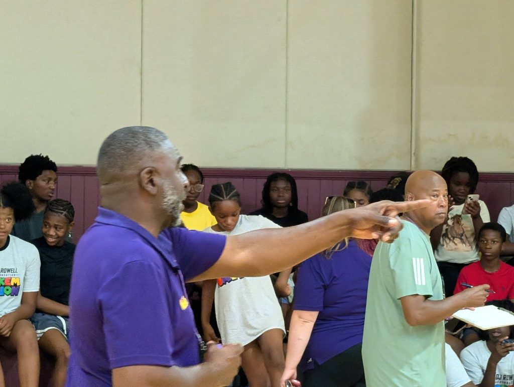 A man in a purple shirt points while another man holds a notebook; a group of seated and standing people watch in the background.
