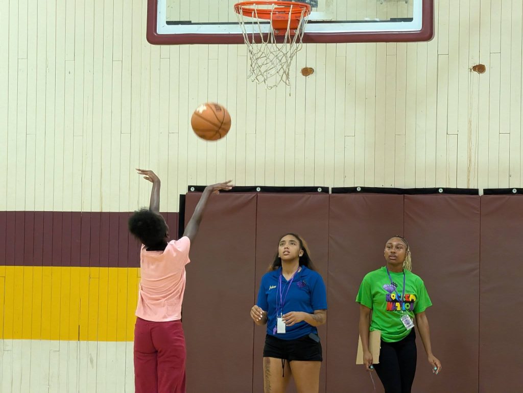 A person shoots a basketball towards the hoop in a gym while two others watch, standing near padded walls.