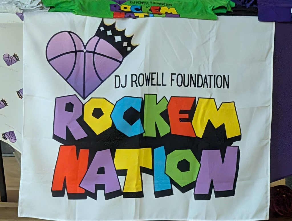 Colorful banner reading "DJ Rowell Foundation Rockem Nation" with a purple heart-shaped basketball and crown graphic above the text.
