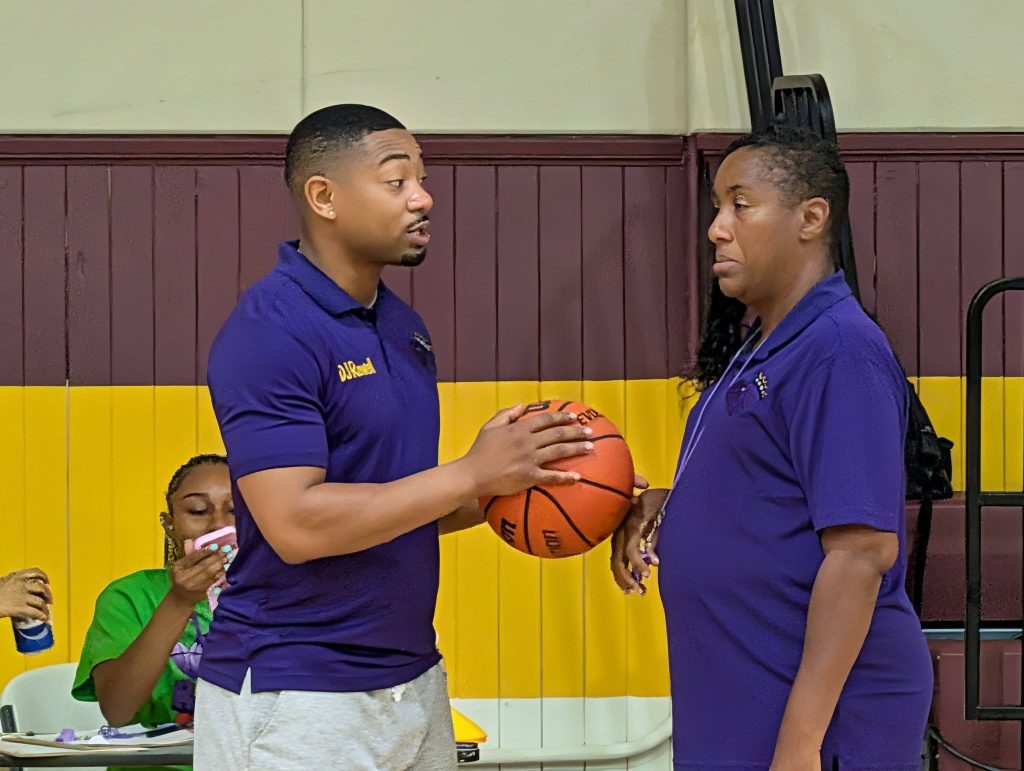 Two coaches in purple shirts are having a serious conversation while holding a basketball inside a gymnasium. Another person sits in the background.