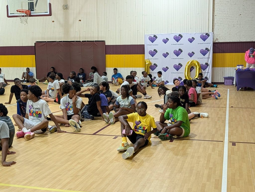 Children sitting on a gym floor in groups, wearing summer camp shirts, with a basketball hoop and a decorated backdrop with balloons in the background.