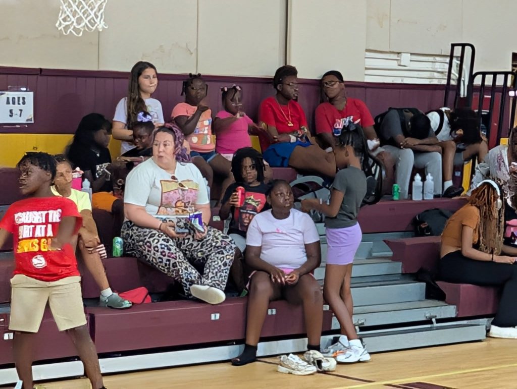 A group of people, including children and adults, sit on bleachers in a gymnasium, some interacting, others watching or resting. A sign reads "AGES 5-7".