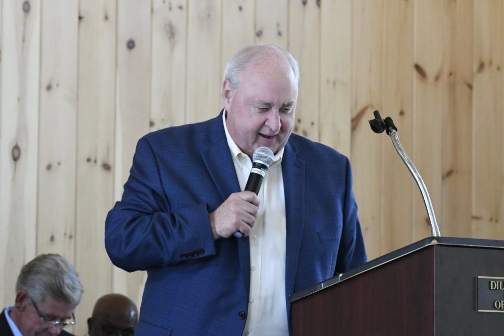 A man in a blue blazer speaks into a microphone at a podium, with wood-paneled walls in the background and other people seated nearby.