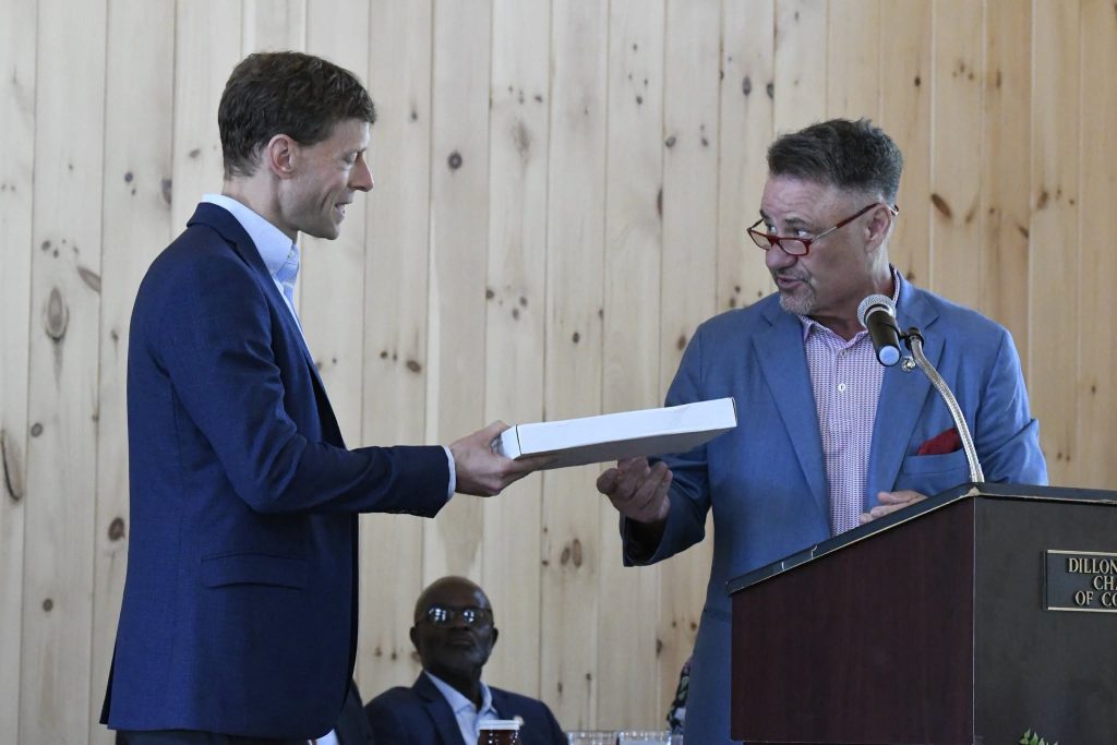 Two men in suits exchange a rectangular box at a podium, with a microphone, in a wood-paneled room. Another person is seated in the background.