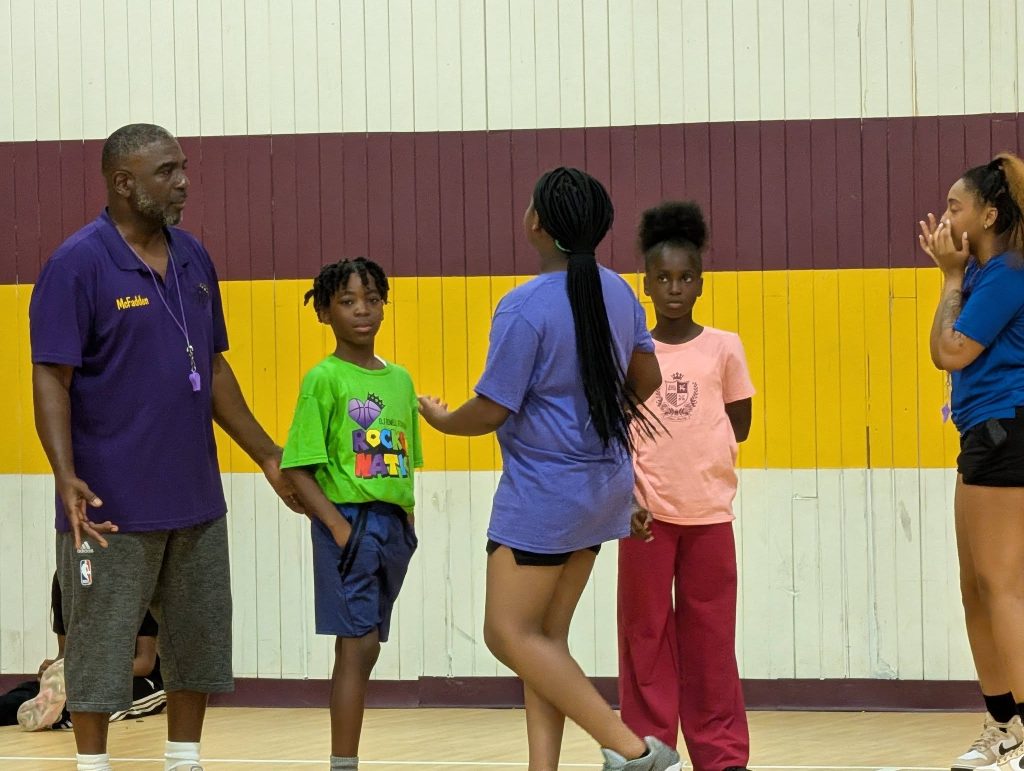 A coach and four children stand together in a gym, having a discussion, with a painted yellow and maroon wall in the background.