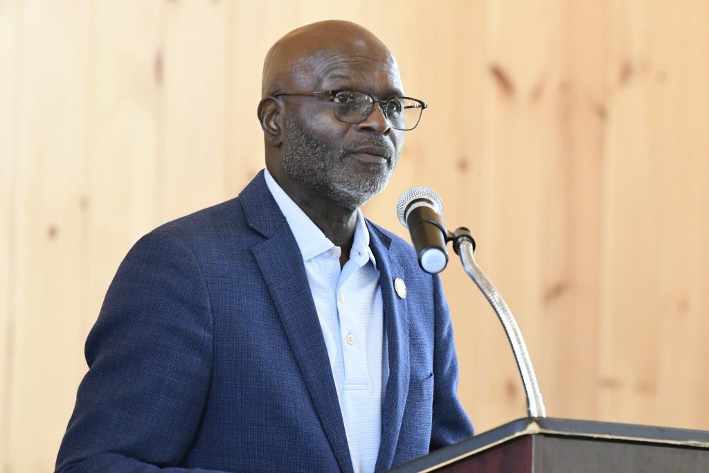 Man in a blue blazer and glasses speaking at a podium with a microphone, wood-paneled background.