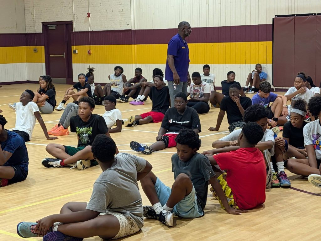 A group of kids sit on a gym floor, listening to an adult instructor standing in the center, during what appears to be a sports or recreational activity.