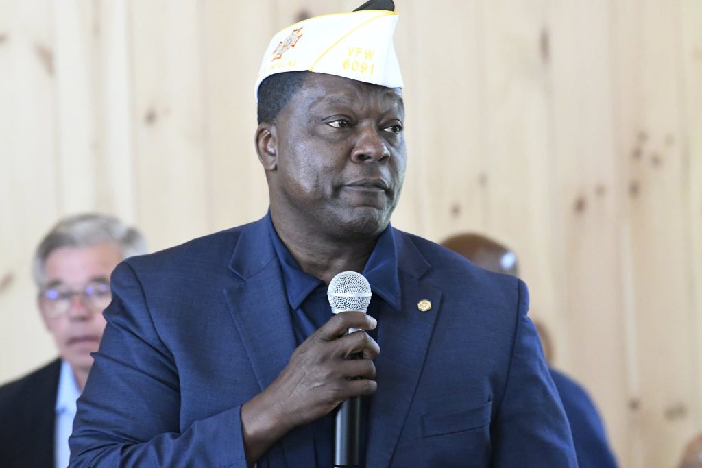 A man in a blue suit and white VFW hat holds a microphone, speaking at an indoor event with people in the background.