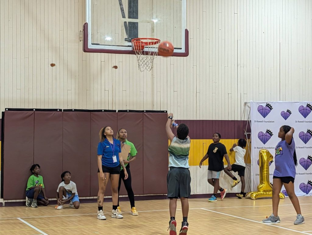 A group of children and teens play basketball in an indoor gym, with one kid shooting the ball toward the hoop while others watch.