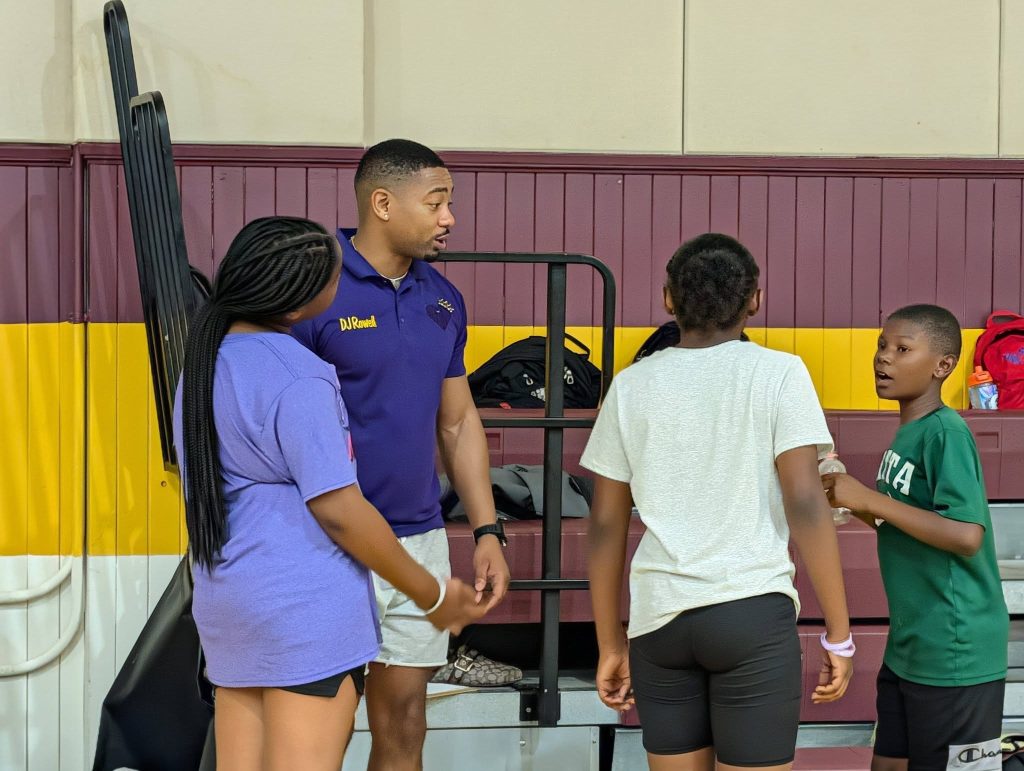 A coach in a purple shirt talks with three children in a gymnasium, standing near bleachers and sports bags.