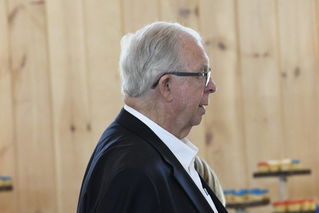 elderly man with gray hair and glasses, wearing a dark jacket and white shirt, stands indoors against a light wooden wall background.