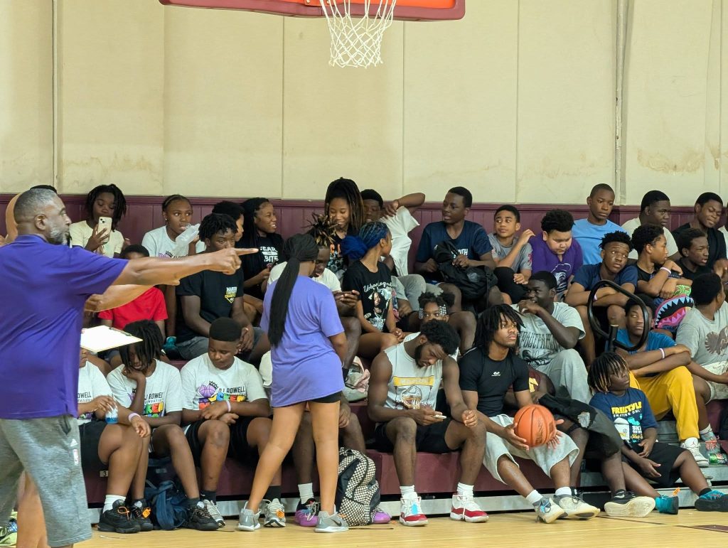 A group of teenagers sit on bleachers in a gym, some using phones, with a coach or instructor pointing and holding a clipboard in the foreground.