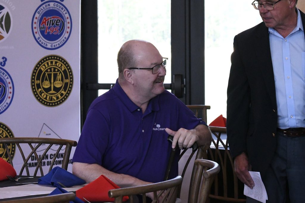 A man in a purple shirt sits at a table with colorful napkins, talking to another man standing beside him at an event.