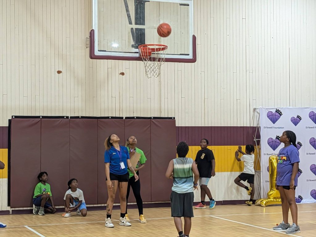 Children and teens play basketball in a gym, watching a ball as it approaches the hoop, with some sitting and some standing on the court.
