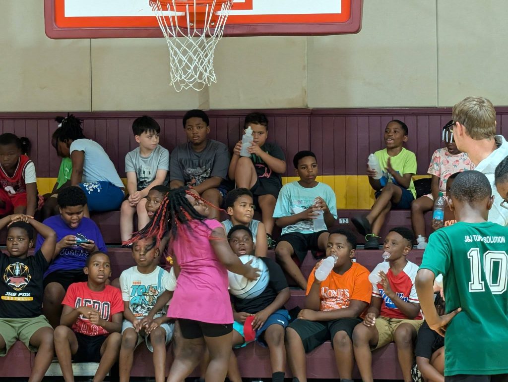 Children sitting on bleachers in a gym, some drinking water, with a basketball hoop above, and a few kids and adults standing in the foreground.