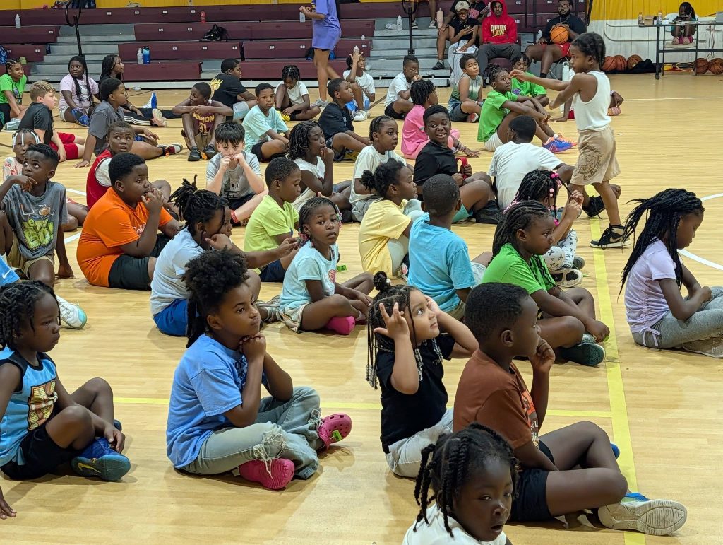 Children sit on a gym floor, attentively listening and interacting, while participating in a group activity or event.