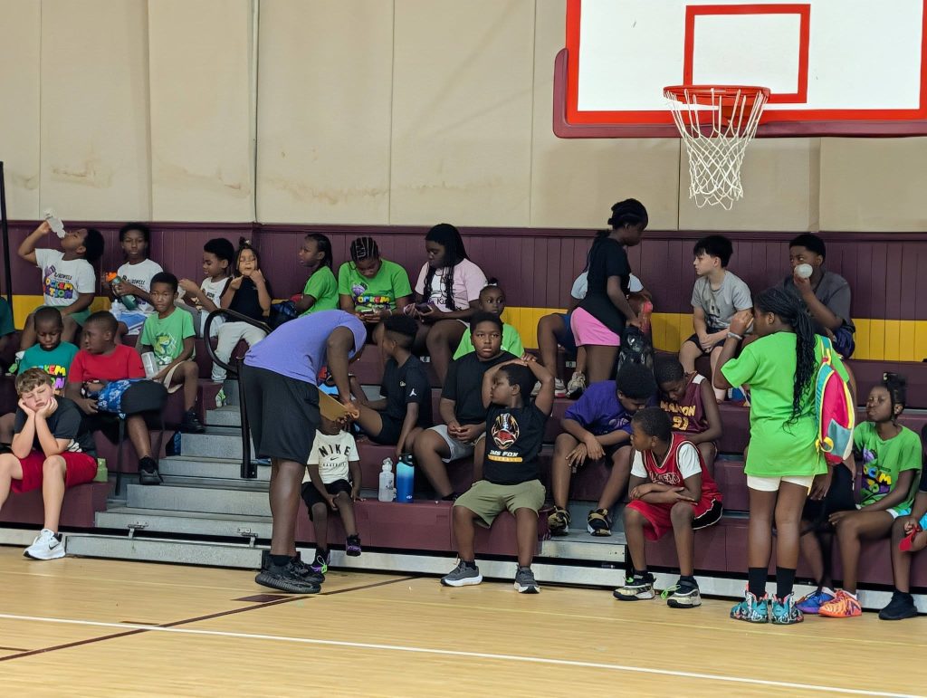 A group of children sit on gym bleachers, some talking, drinking, or eating, while others stand nearby under a basketball hoop.