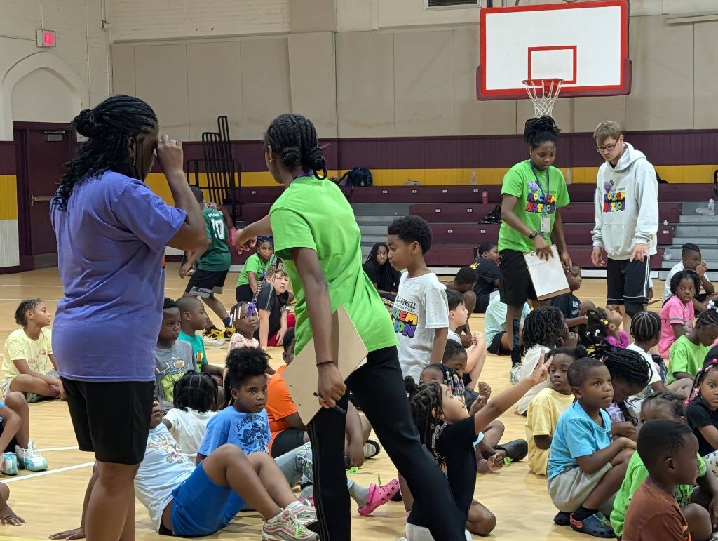 Adults and children gathered in a gymnasium; adults are supervising and talking while children sit on the floor, appearing to participate in a group activity or camp event.