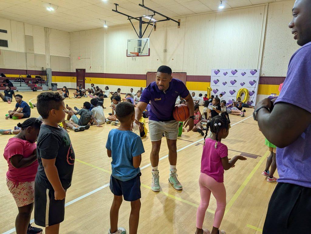 A coach interacts with children in a gymnasium during a basketball event, while more kids and adults sit and gather in the background.