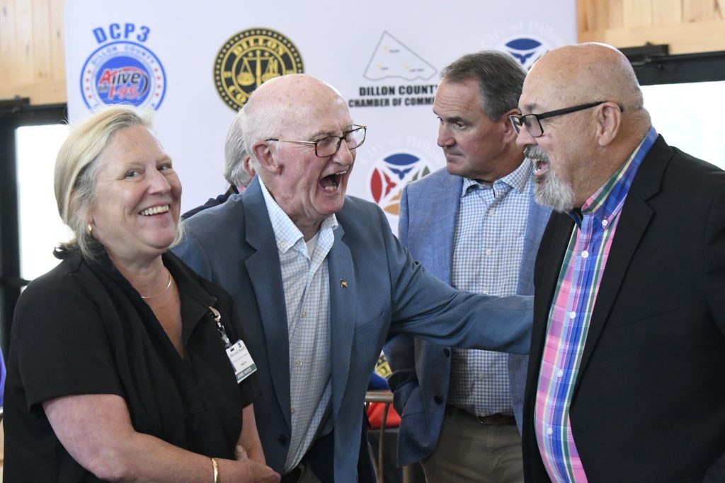 Four adults in business attire stand together, smiling and laughing, with event banners visible in the background.