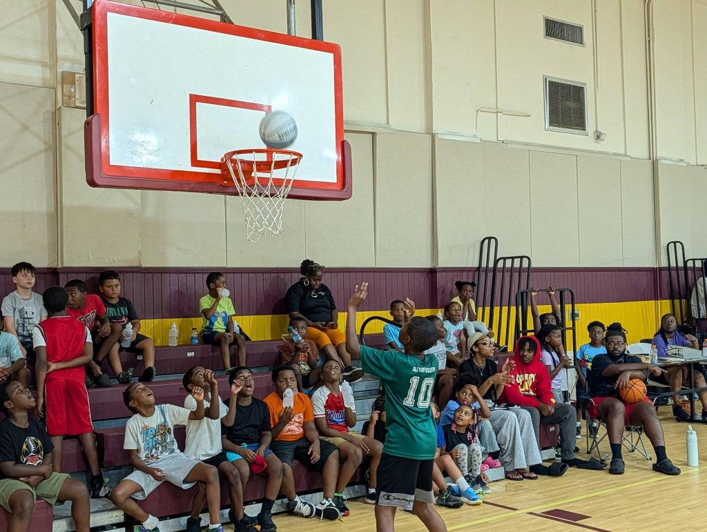 A boy shooting a basketball in a gym as a group of kids and adults watch from the bleachers.