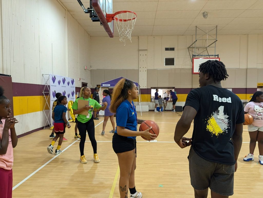 Children and adults gather in a gymnasium, some holding basketballs, while a coach instructs near the basketball hoop during a recreational or training event.