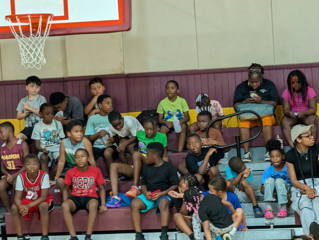 A group of children and a few adults sit on gym bleachers under a basketball hoop, some talking, eating, or looking at their phones.