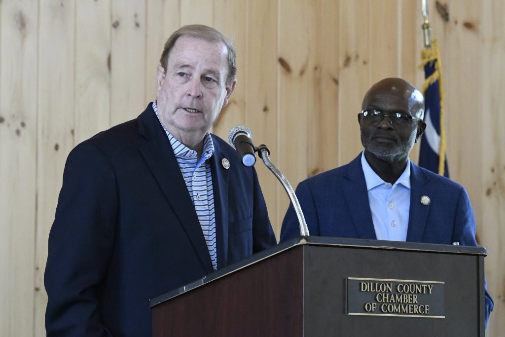 Two men in suits stand at a podium labeled "Dillon County Chamber of Commerce" during an indoor event with wood-paneled walls.