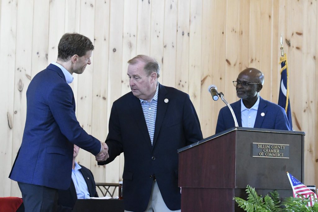Three men in suits interact at a podium labeled "Dillon County Chamber of Commerce" during an indoor event, with one man shaking hands and another standing by the microphone.