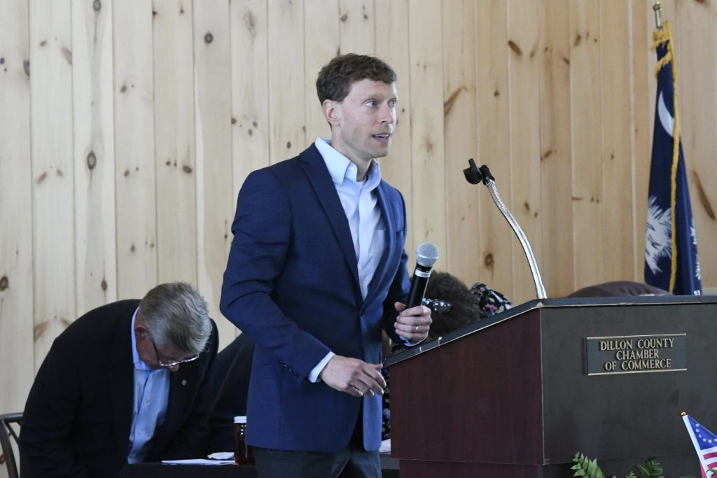 A man in a blue suit speaks into a microphone at a podium labeled "Dillon County Chamber of Commerce," with an American flag visible in the background.
